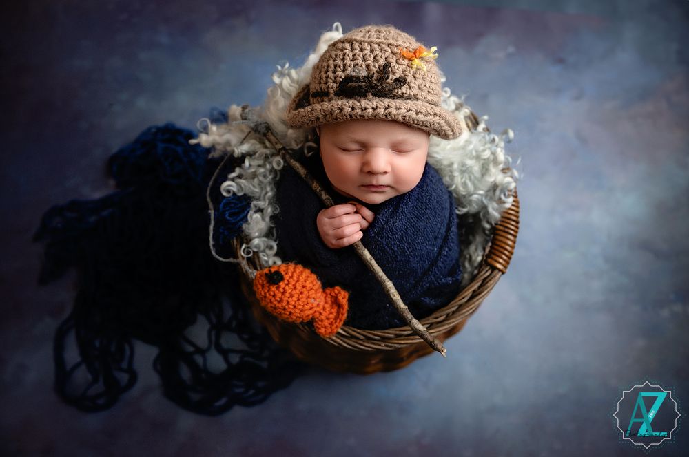 Newborn in fishing attire sleeping in rustic basket with orange prop fish.