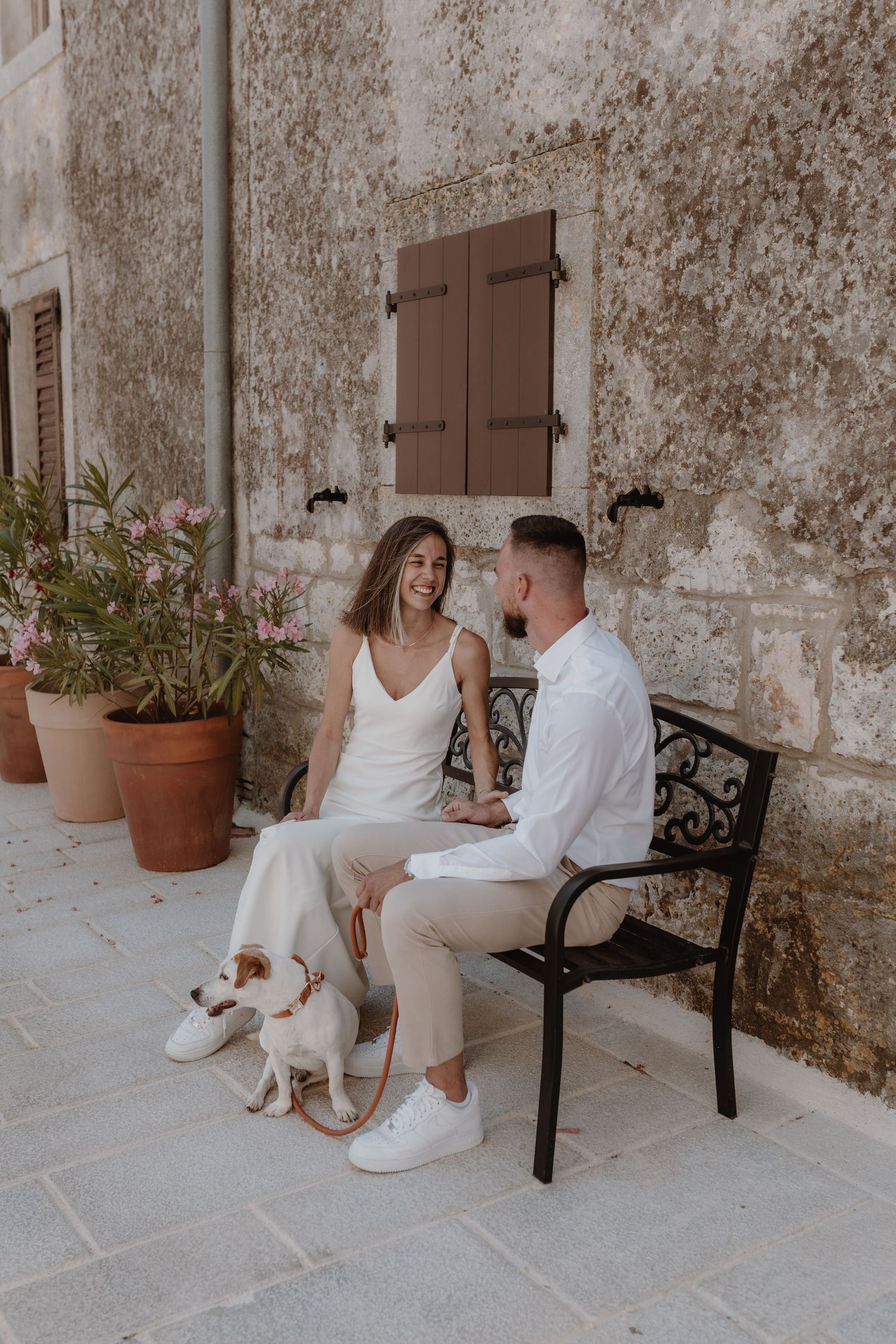 A joyful couple sits together on a concrete bench, the bride wearing an intricate lace gown and the groom in a classic tuxedo.