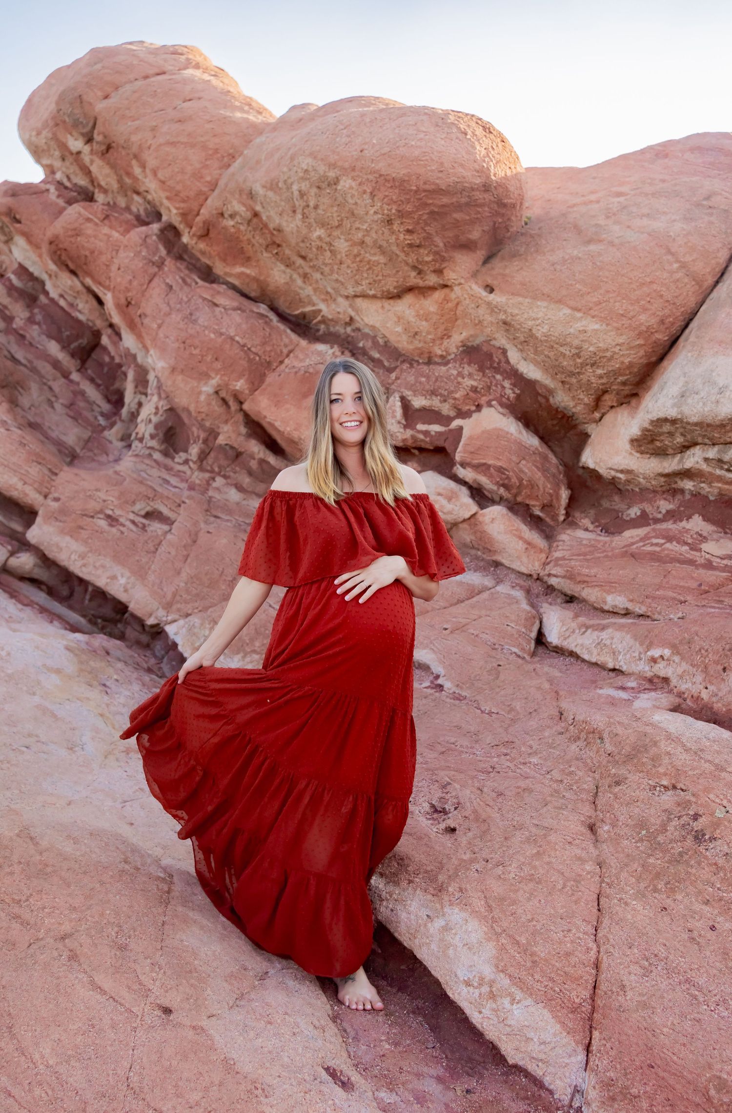 Dramatic maternity portrait in red dress against red rock formations at sunset.
