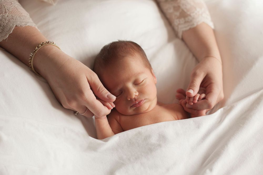 neonato dorme sul bean bag, coperta di cotone bianco. Le mani della mamma lo tengono per le sue piccole manine, sessione fotografica newborn
