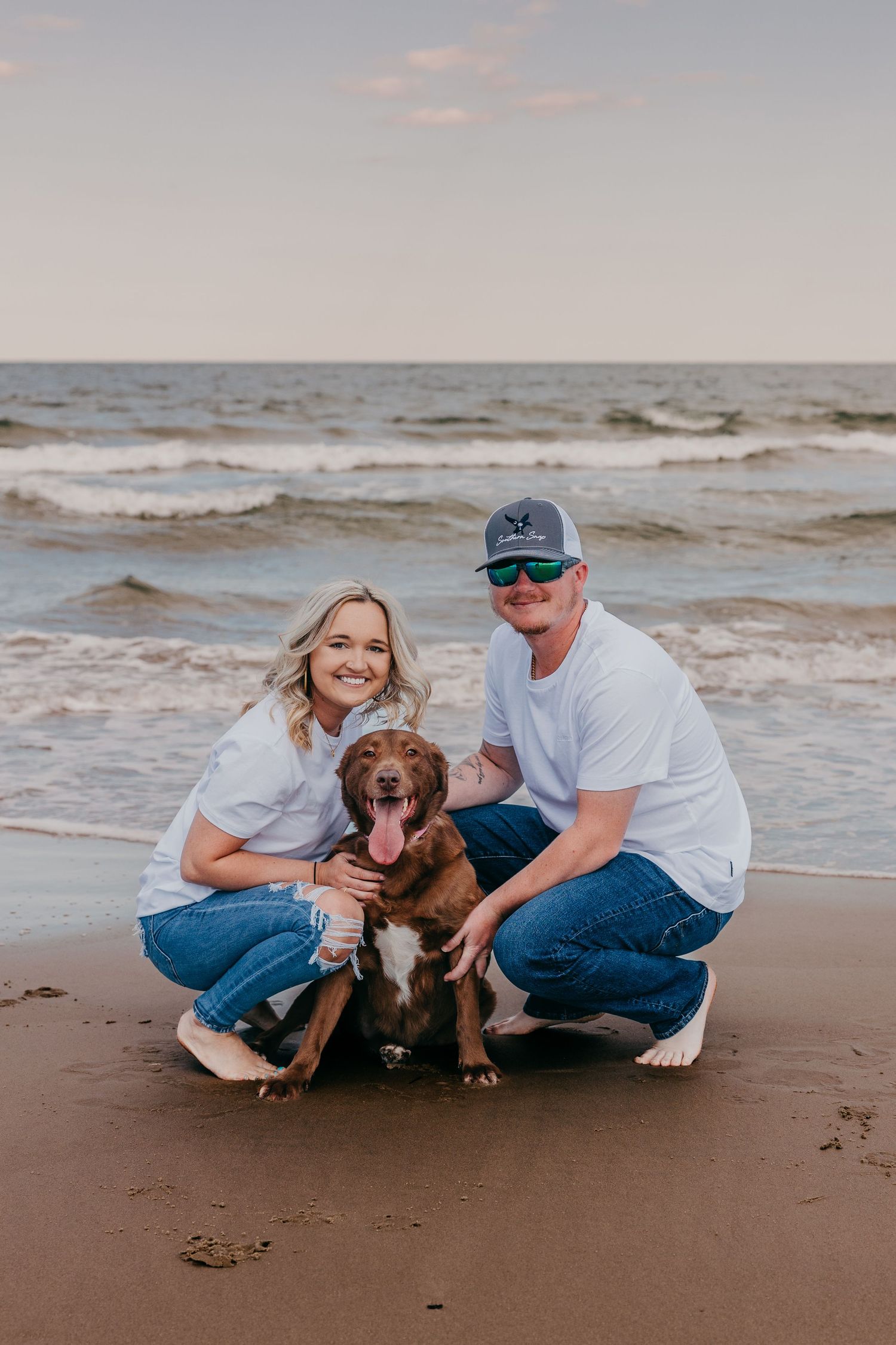 Couple sitting on beach with their brown dog during sunset.