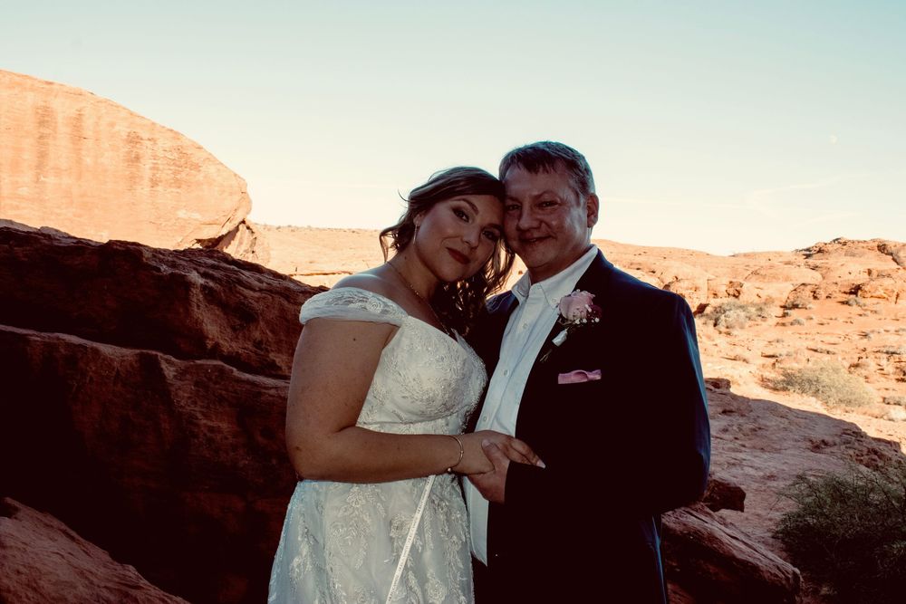 Wedding couple embracing at red rock canyon at sunset, bride in lace dress and groom in dark suit.