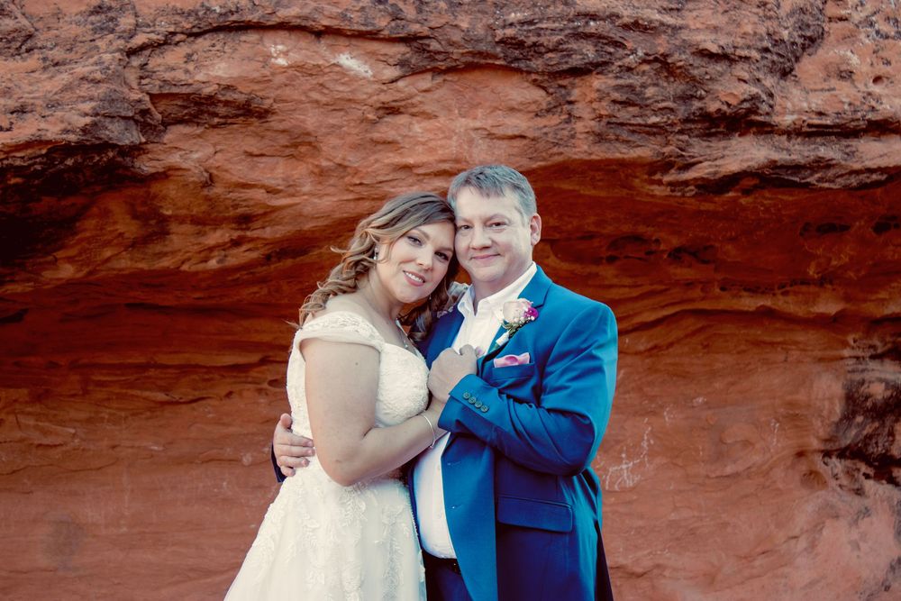 Smiling wedding couple posing together against vibrant red sandstone rock formation outdoors.