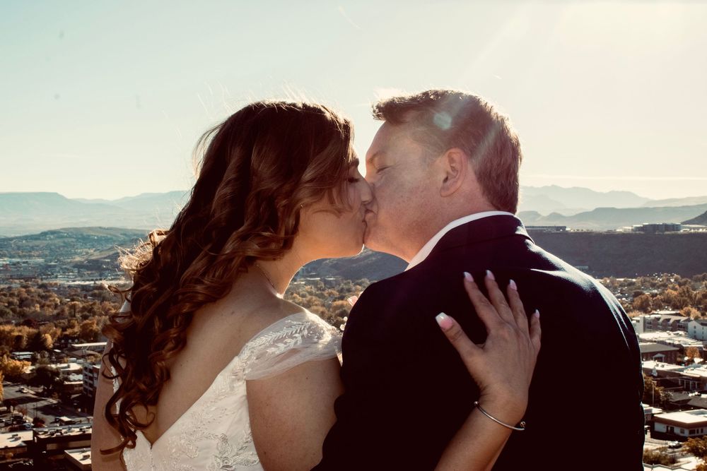 Bride and groom sharing romantic kiss with scenic mountain valley backdrop in sunlight.