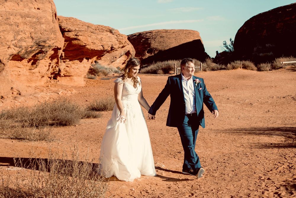 Newlyweds walk hand in hand through red desert landscape in wedding attire.