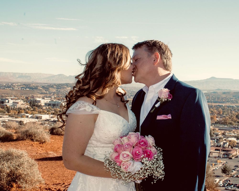 Bride and groom share first kiss on red rock overlook with city views and pink bouquet.