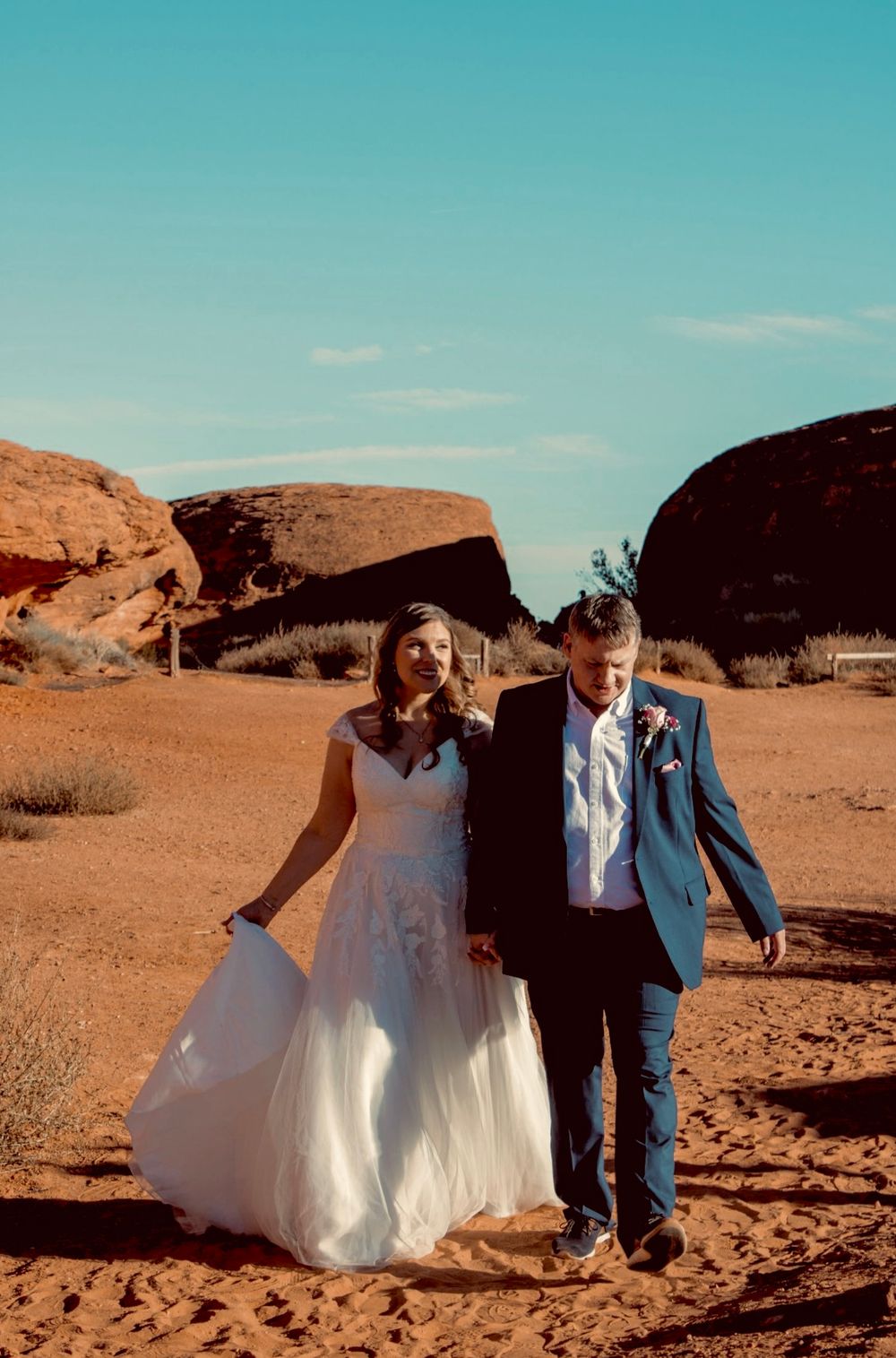 Newlywed couple walks hand-in-hand through red sand desert landscape under bright blue sky.