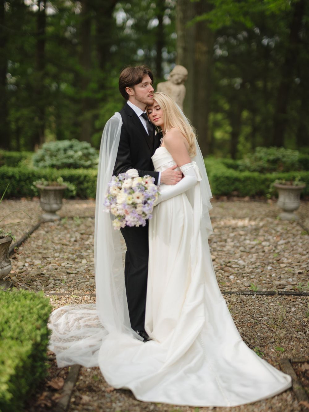 Wedding couple poses in garden setting with bride holding pastel bouquet.