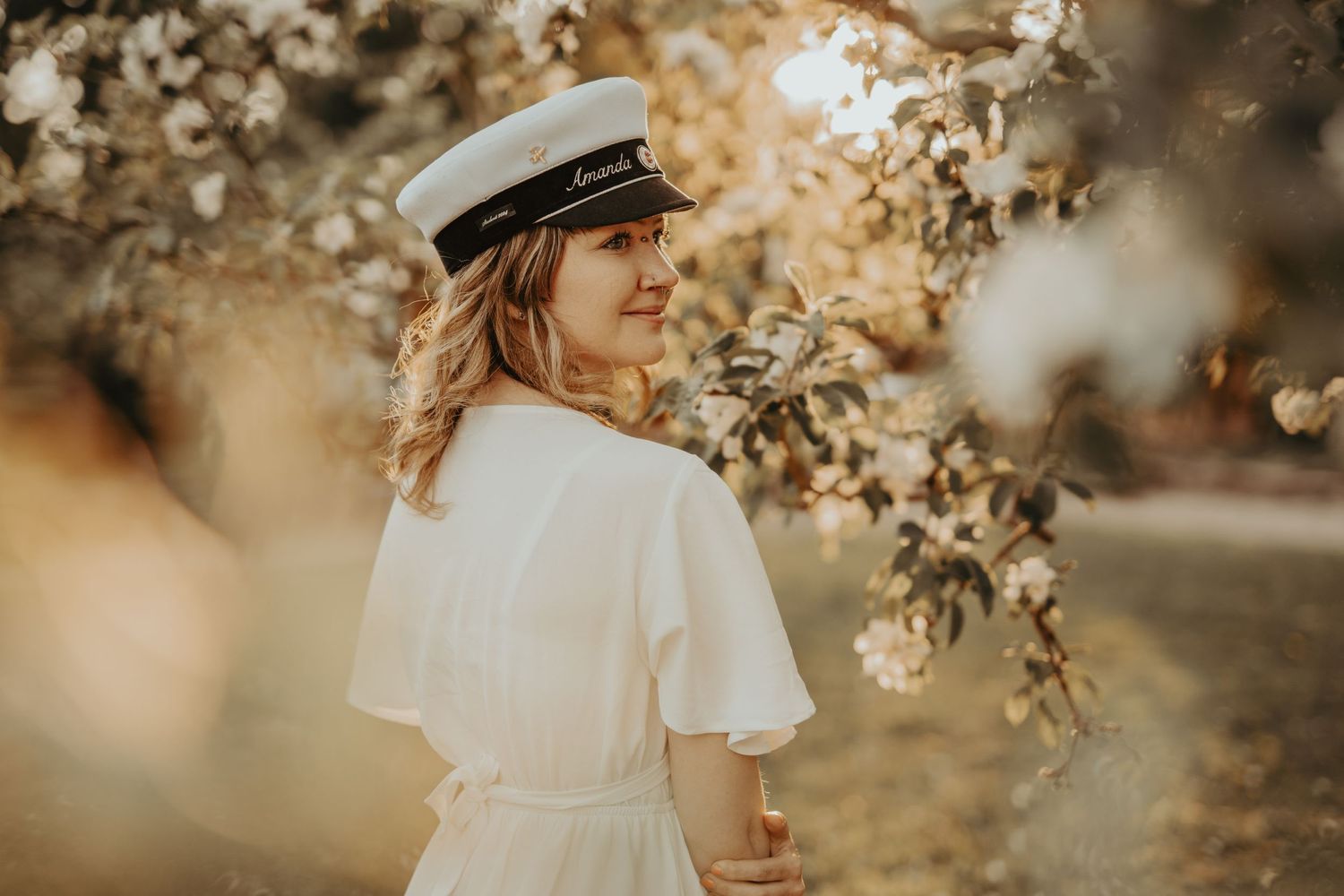 Woman in white dress and nautical cap poses beneath flowering tree branches.