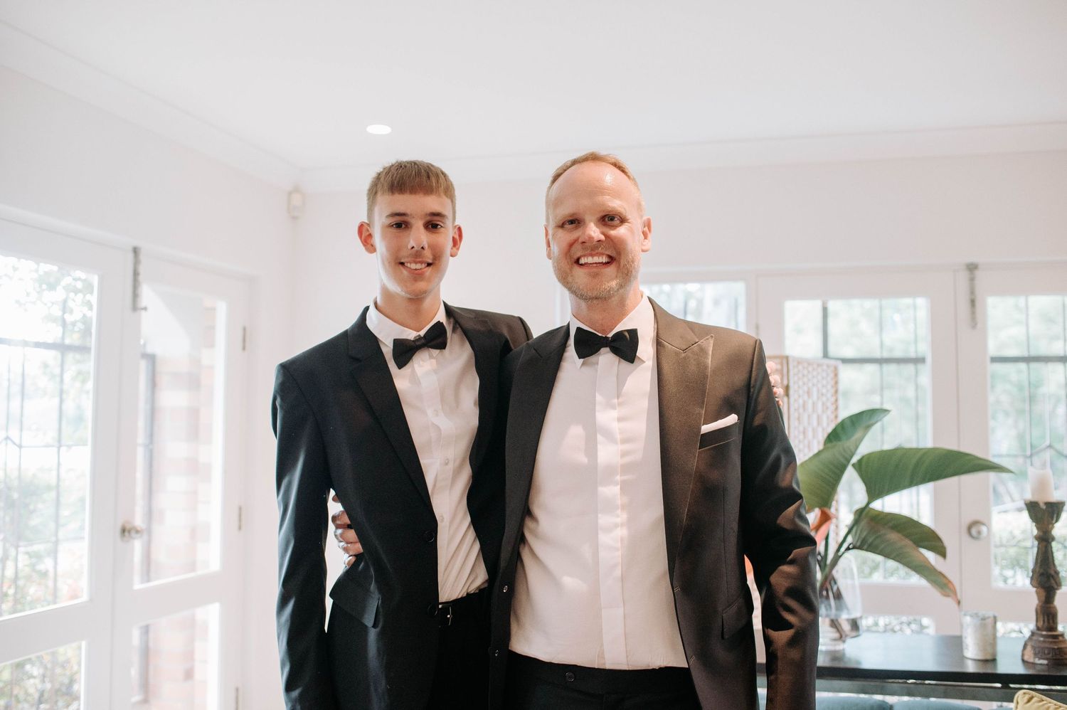 Two people in black tuxedos pose together in a bright white interior space.