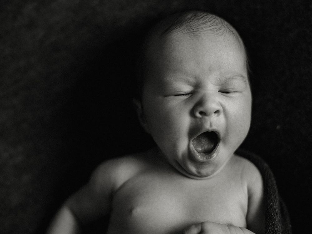 Black and white portrait of yawning newborn baby against dark background.