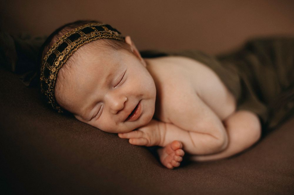 Peaceful newborn wearing decorative headband sleeping on brown backdrop.