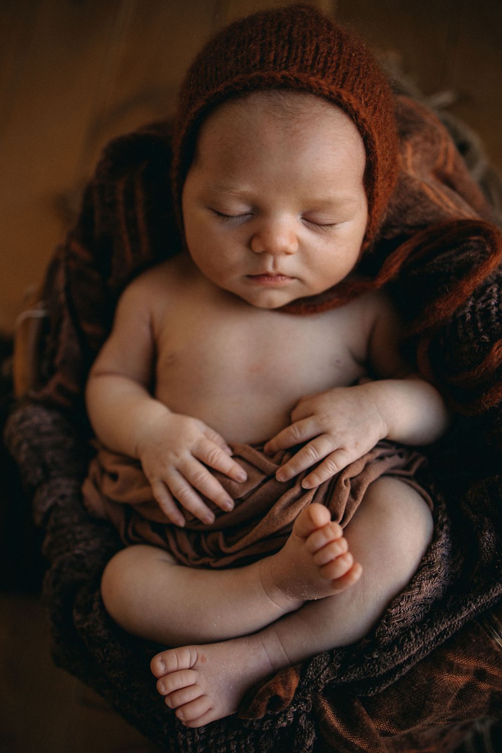 Newborn wearing brown knit hat rests on dark textured fabric during portrait session.