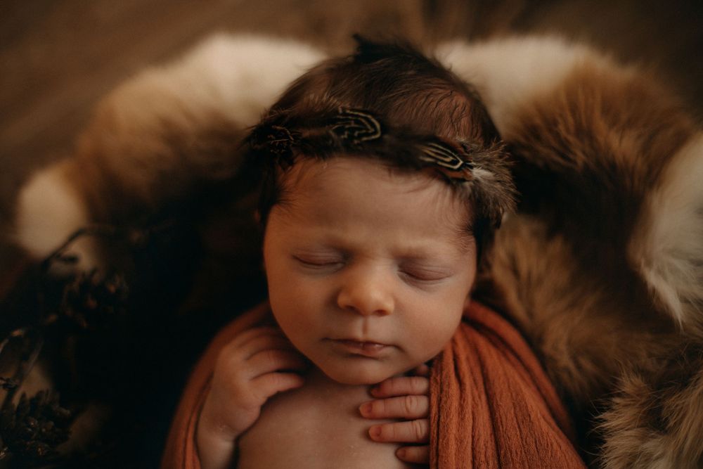 Close-up portrait of sleeping newborn with floral crown and orange wrap on fur background.