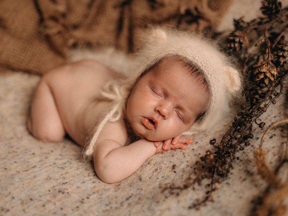Sleeping newborn wearing white bonnet photographed with dried flowers on beige backdrop.