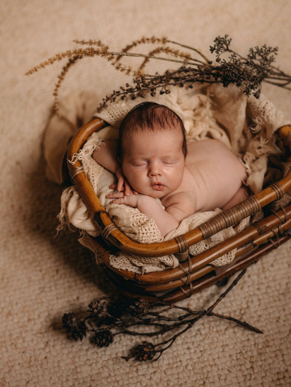 Newborn sleeping peacefully in wicker basket with dried floral decorations.