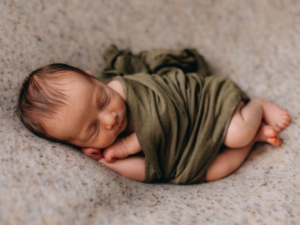 Sleeping newborn wrapped in olive green fabric on textured carpet.
