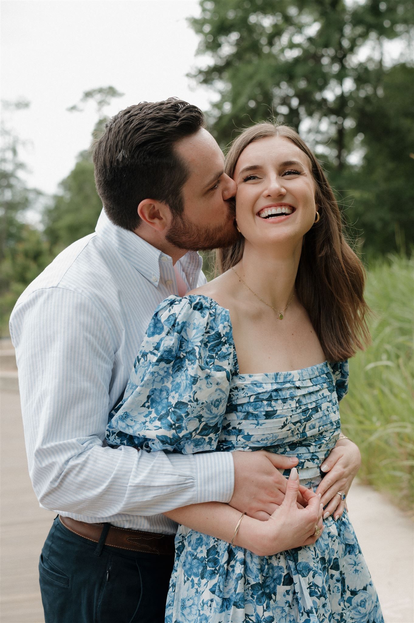 A romantic outdoor portrait featuring a couple embracing, with her wearing a blue floral dress in a natural setting.