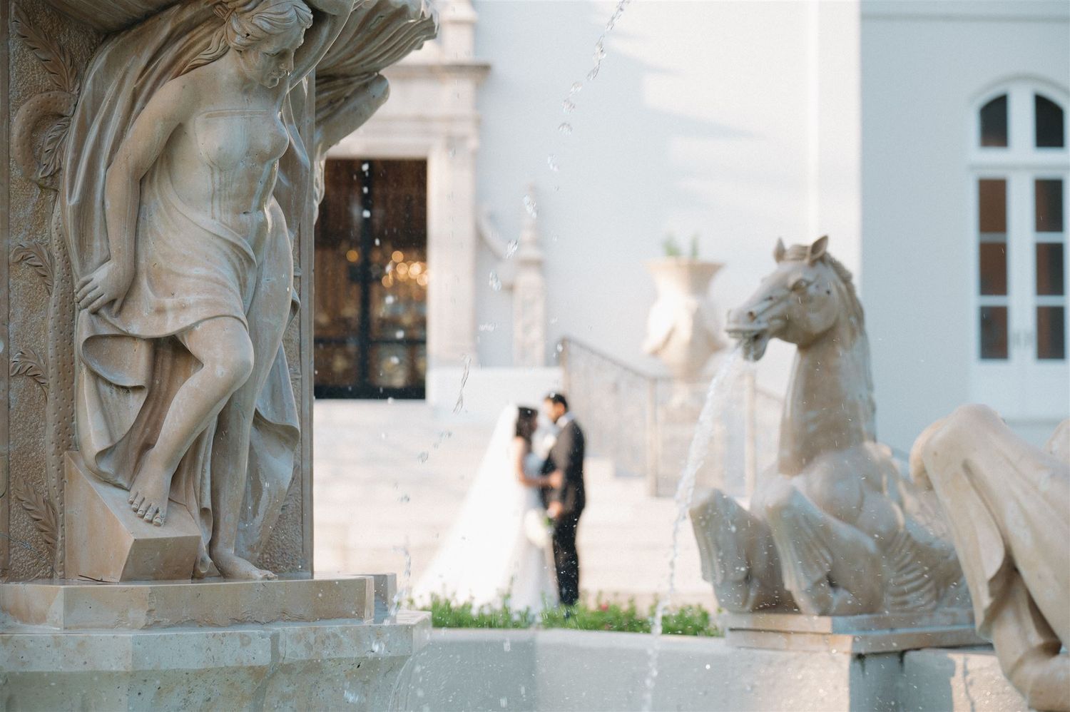 Classical stone statues frame a distant wedding couple at an elegant white venue.