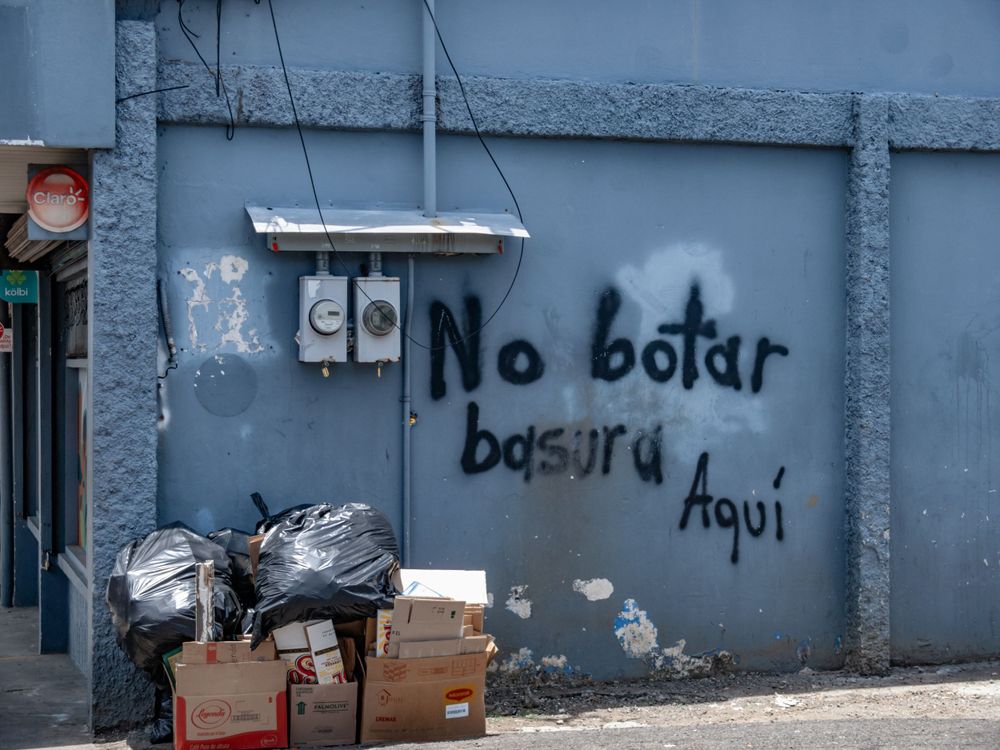 Graffiti reading 'No botar basura aqui' on blue wall with trash bags and boxes on ground.