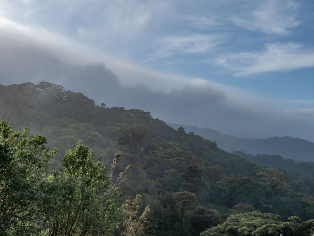 Dramatic mountain landscape with fog rolling over green hills under a blue sky.