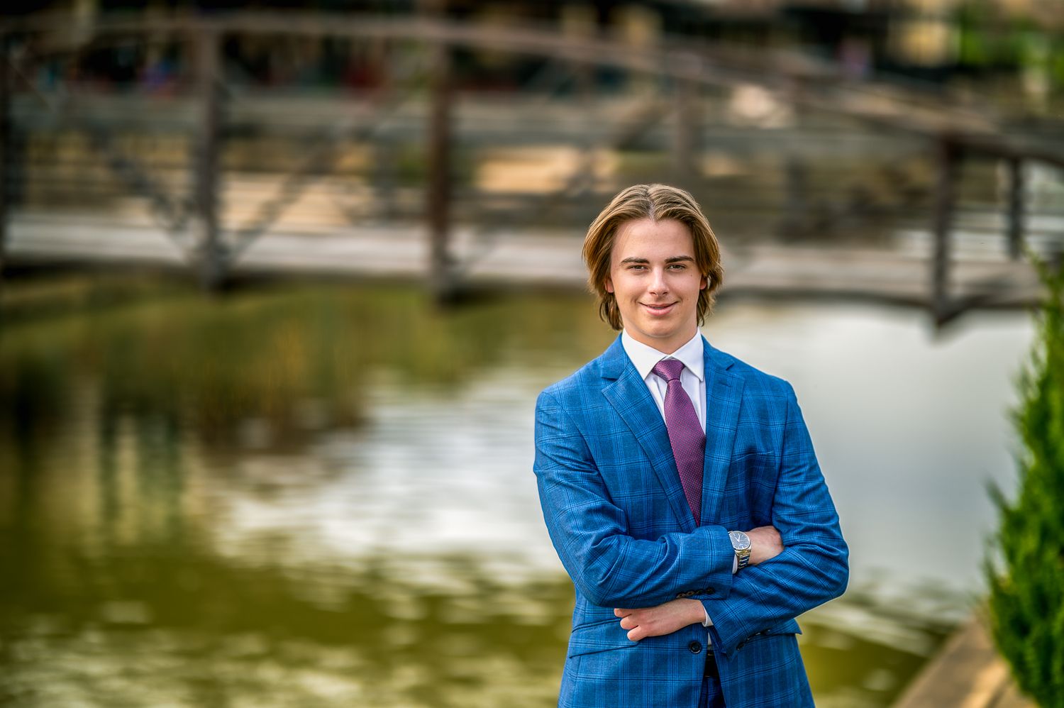 A person in a blue suit standing confidently in front of a pond with a bridge in the background.