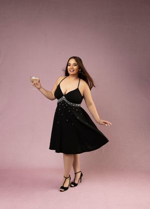 Woman in sparkly black cocktail dress holds champagne glass while posing against pink background.