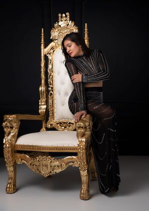 Dramatic portrait on ornate gold throne chair wearing black embellished outfit.