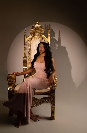 Woman in pink gown sits elegantly on ornate golden throne with dramatic lighting and shadows.