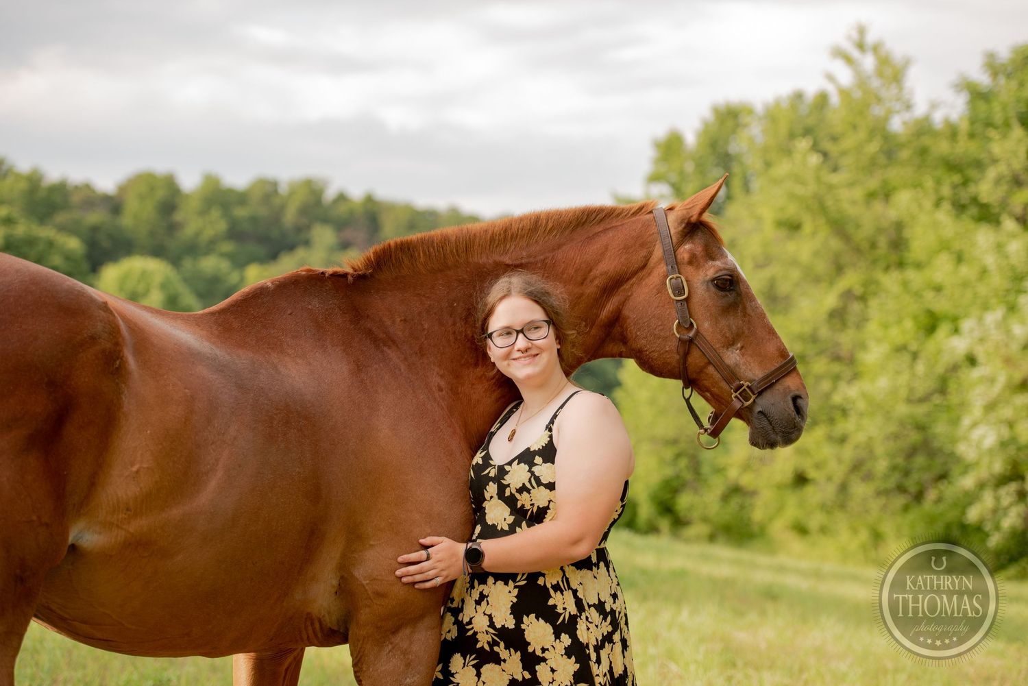 A chestnut horse stands next to someone in a floral dress in a sunny pasture with trees in the background.