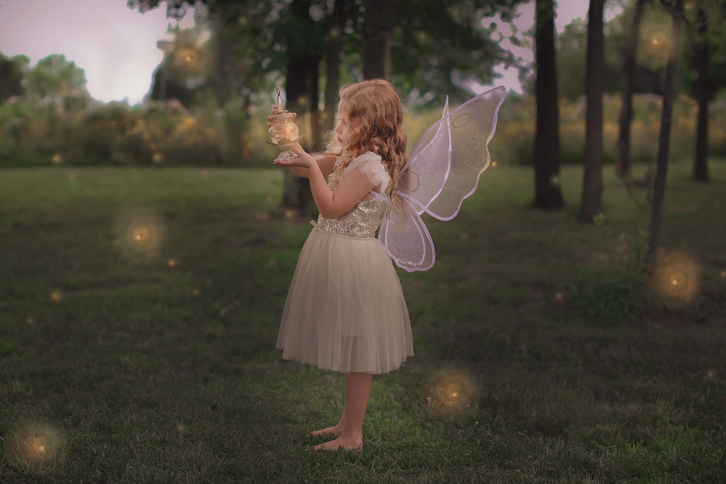 Young child in white dress and fairy wings plays with glowing lights in garden setting.