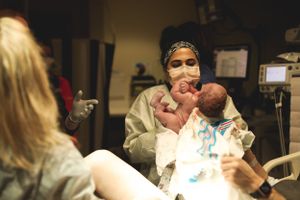 Healthcare worker in protective gear holds newborn baby in hospital delivery room.