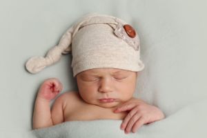 Sleeping newborn baby wearing a beige knit cap rests peacefully against a soft backdrop.