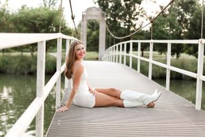 Fashion portrait of a woman in white dress sitting on wooden bridge over lake at sunset.
