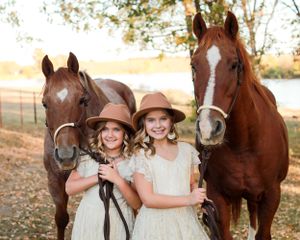Two young girls in vintage dresses pose with two brown horses in an autumn setting.