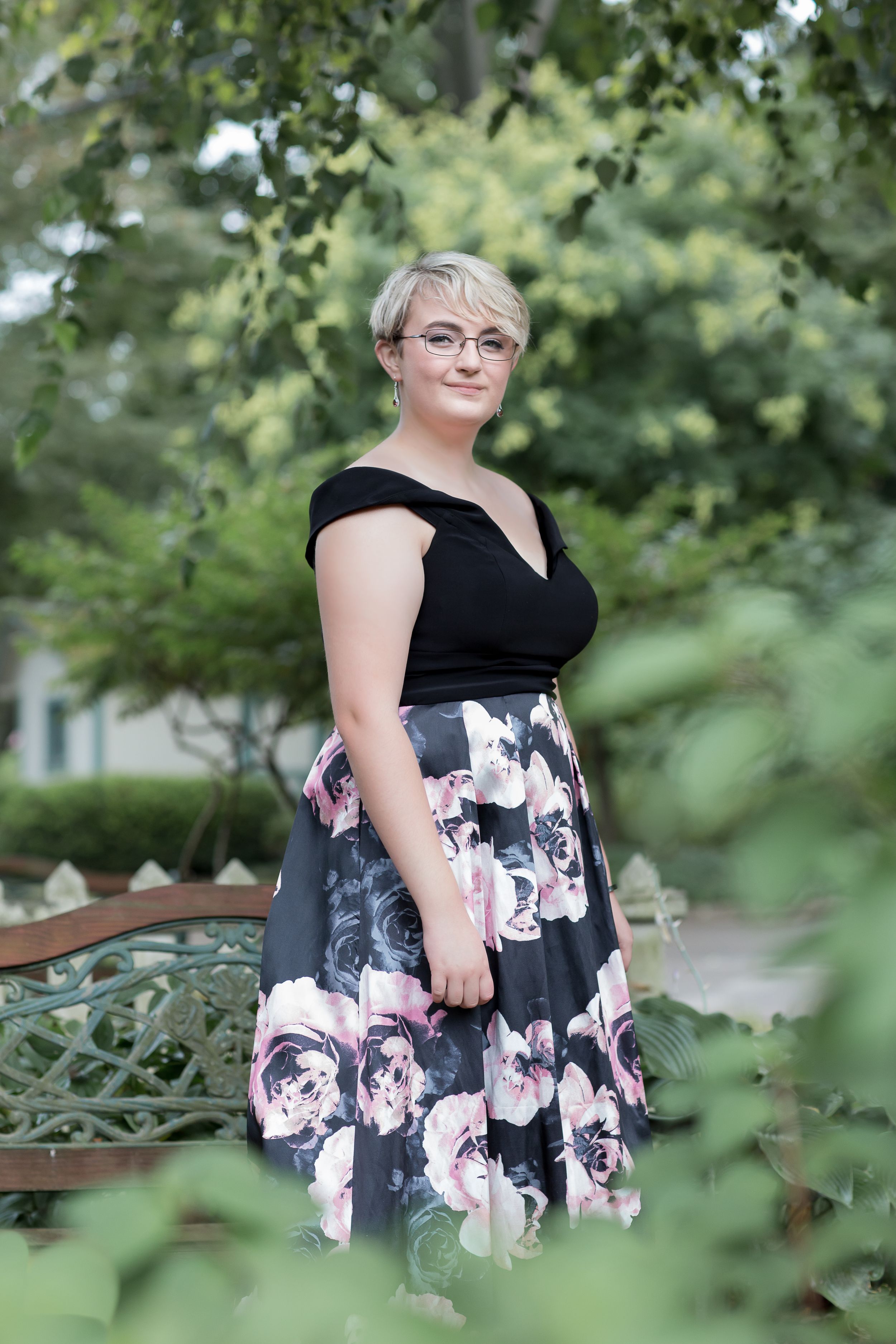 Woman with short blonde hair in floral skirt posing in lush garden setting.