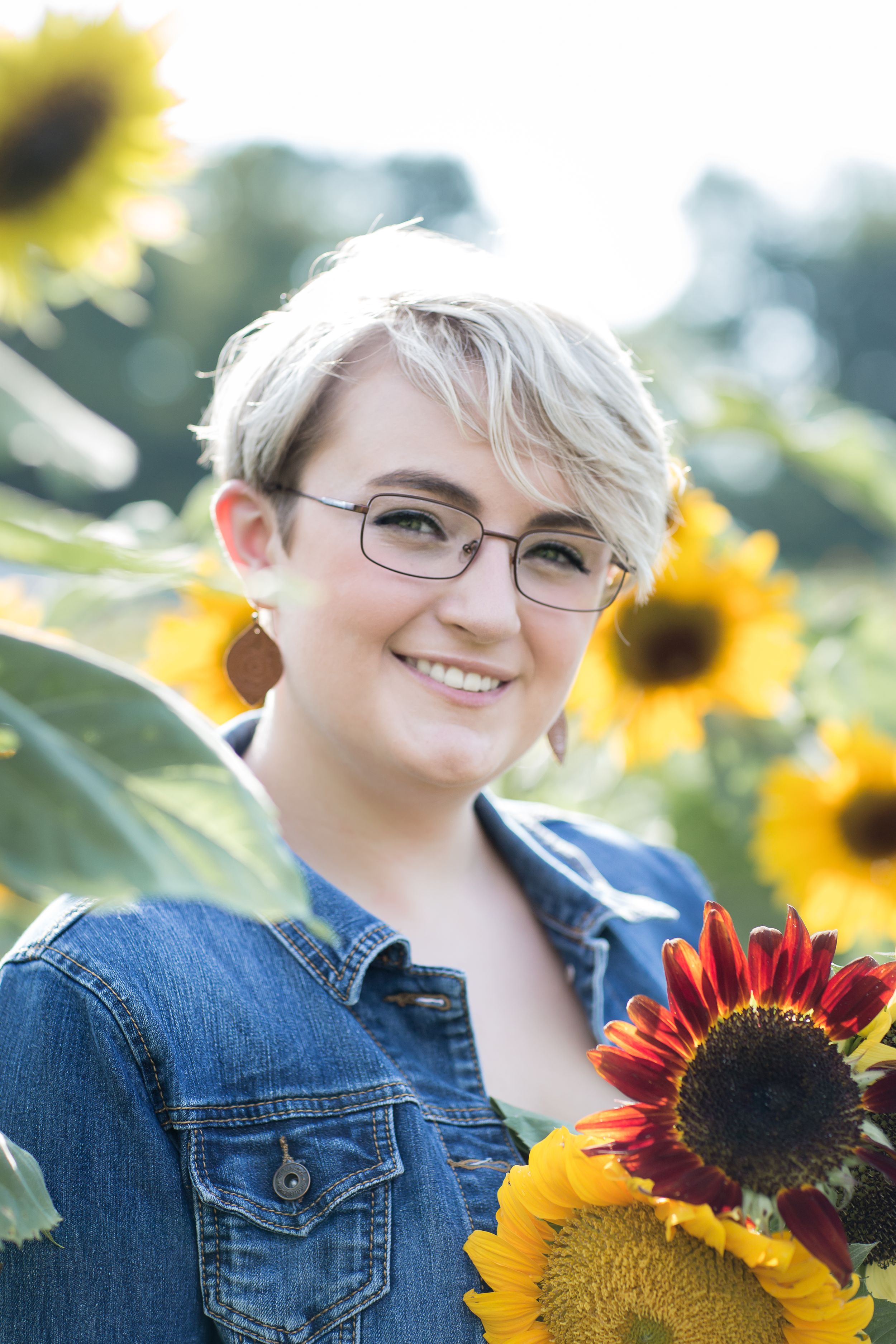 Smiling person with short blonde hair holding sunflowers in bright sunflower field.