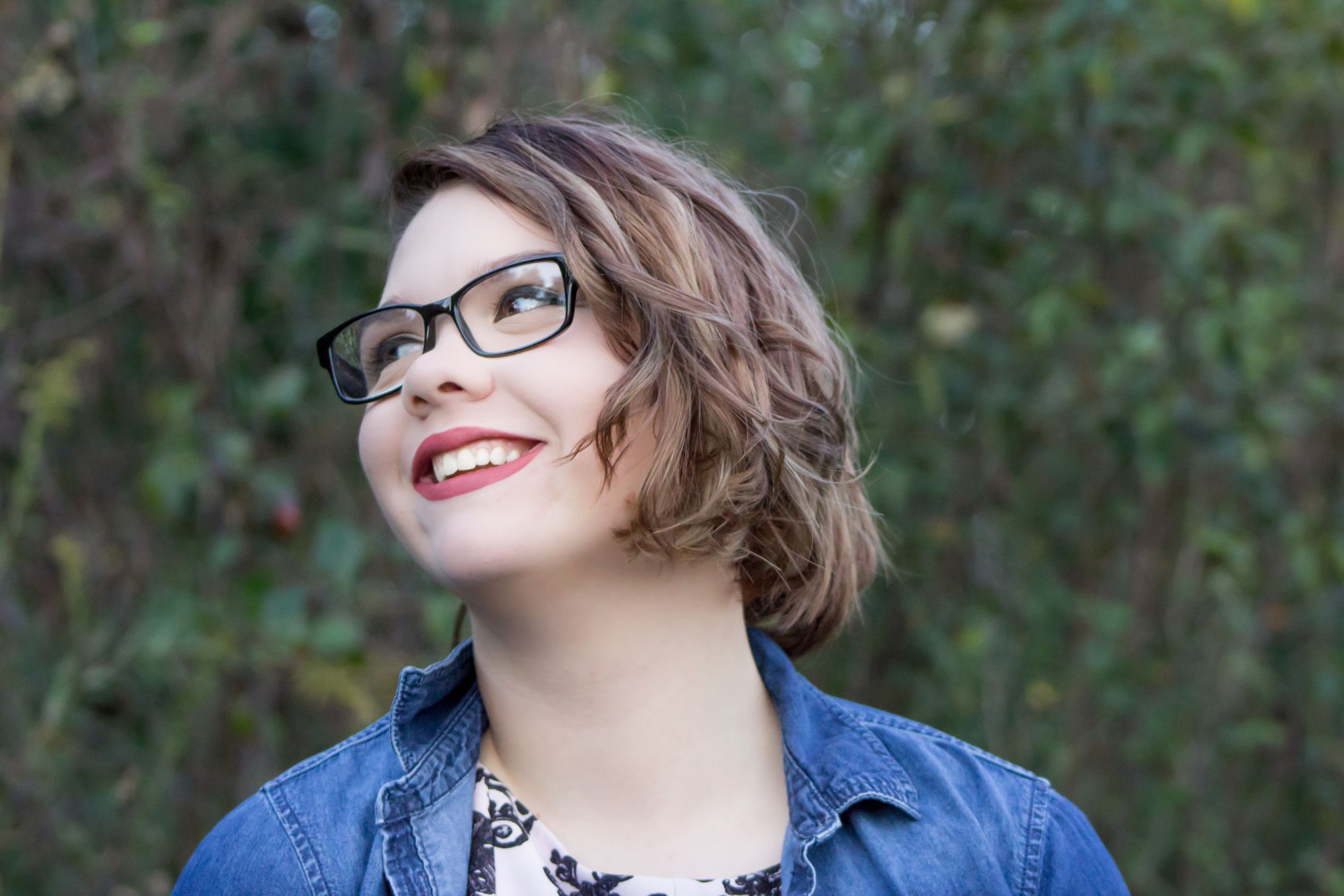Smiling woman with glasses and red lips looking up outdoors near green foliage.