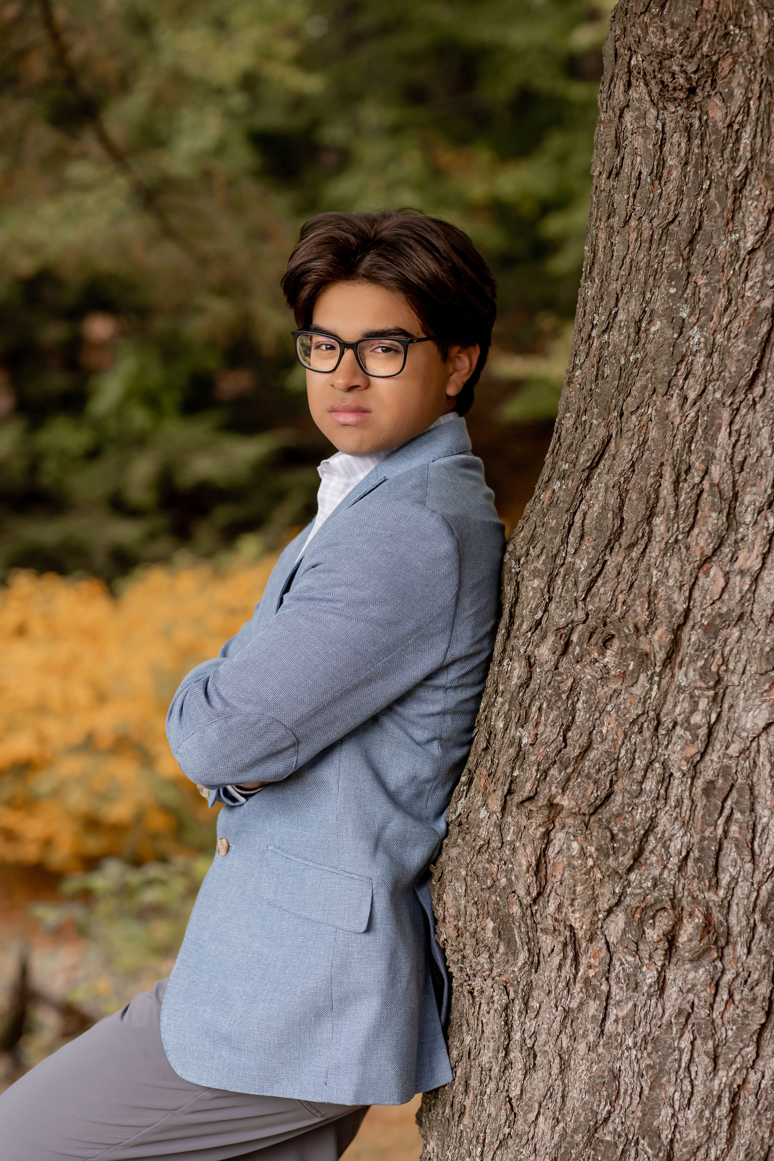 Young man in gray blazer leaning against tree trunk in autumn park setting.