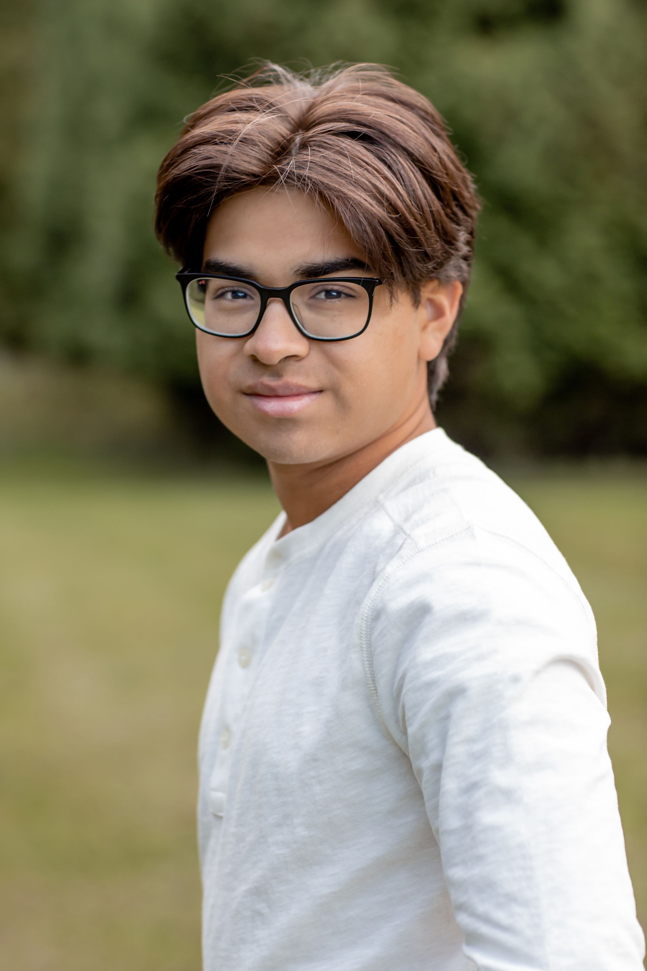 Young man with glasses in white shirt smiling outdoors in green park setting.