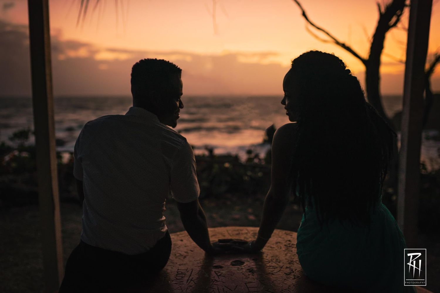 Silhouette of a couple sitting together watching a romantic beach sunset through palm trees.