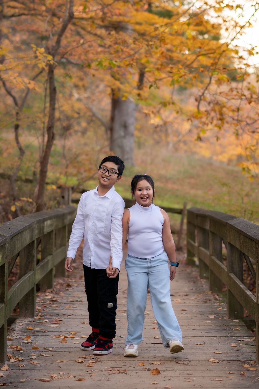 Two children holding hands walking on a wooden bridge surrounded by colorful autumn foliage.monmouth county, Homdel Park NJ