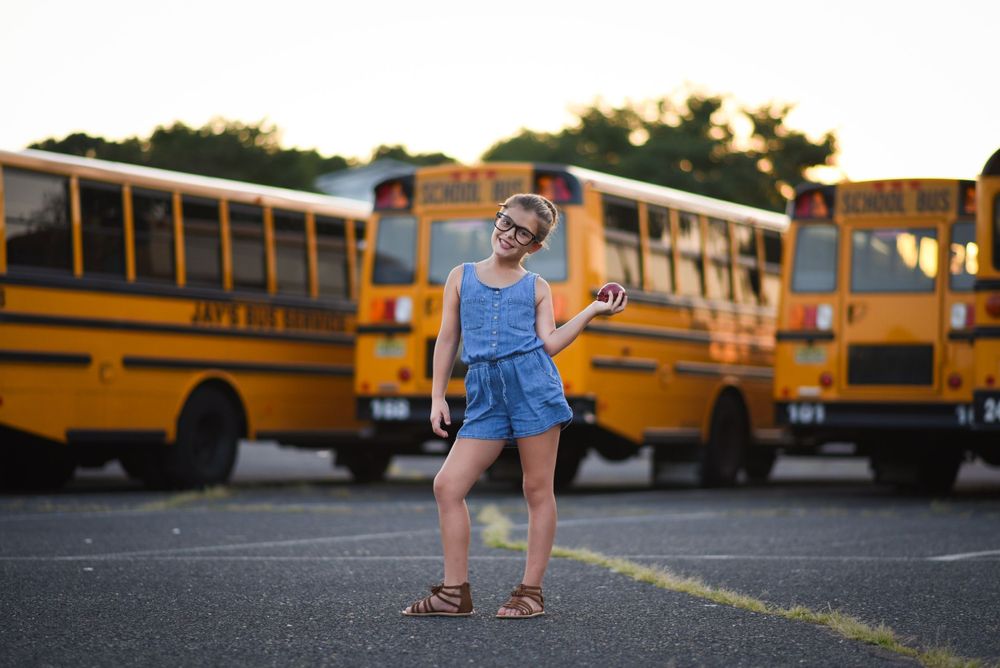 Child in denim romper in front of yellow school buses at sunset, holding an apple. Back to school portrait.Middletown NJ