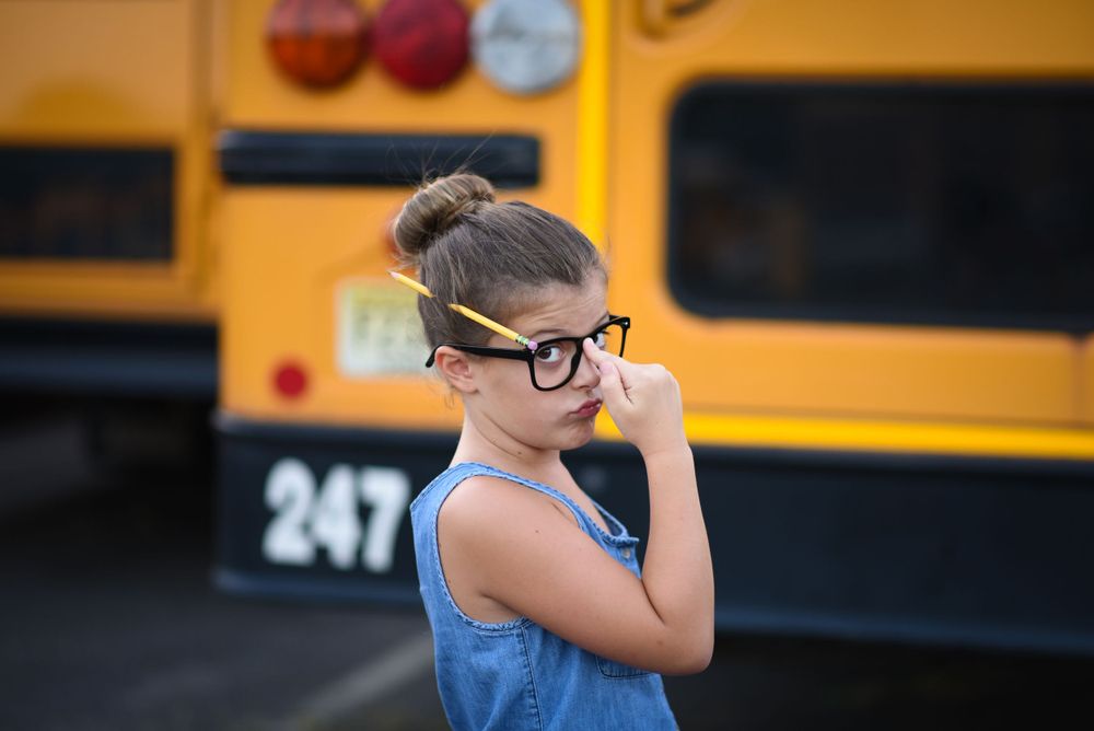A stylish pose of young school girl in denim and glasses with hair in bun and a yellow pencil in it,  by a yellow school bus. Back to school
