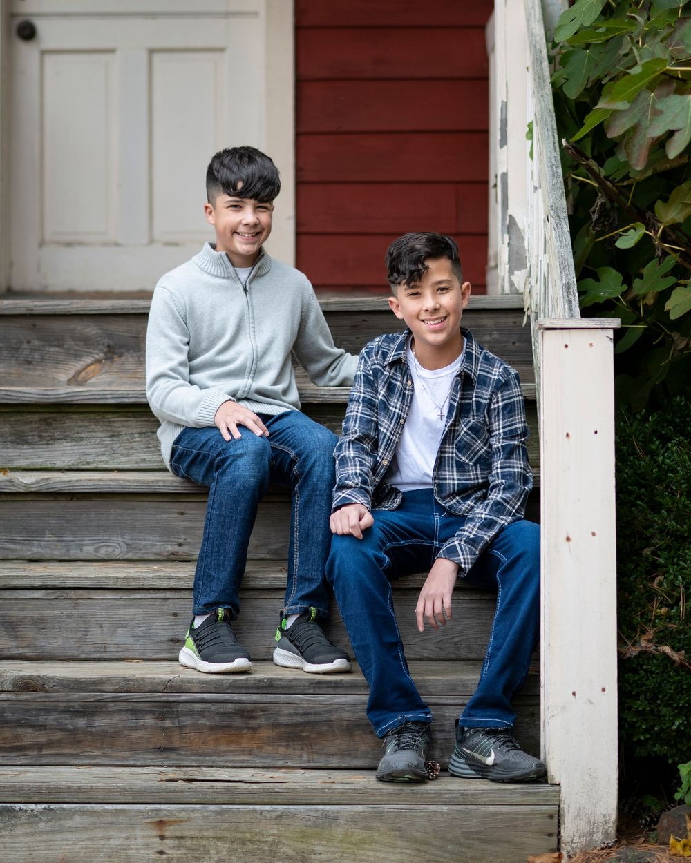 Two teenage boys sitting on rustic wooden porch steps, smiling at camera outside red house.Middletown NJ