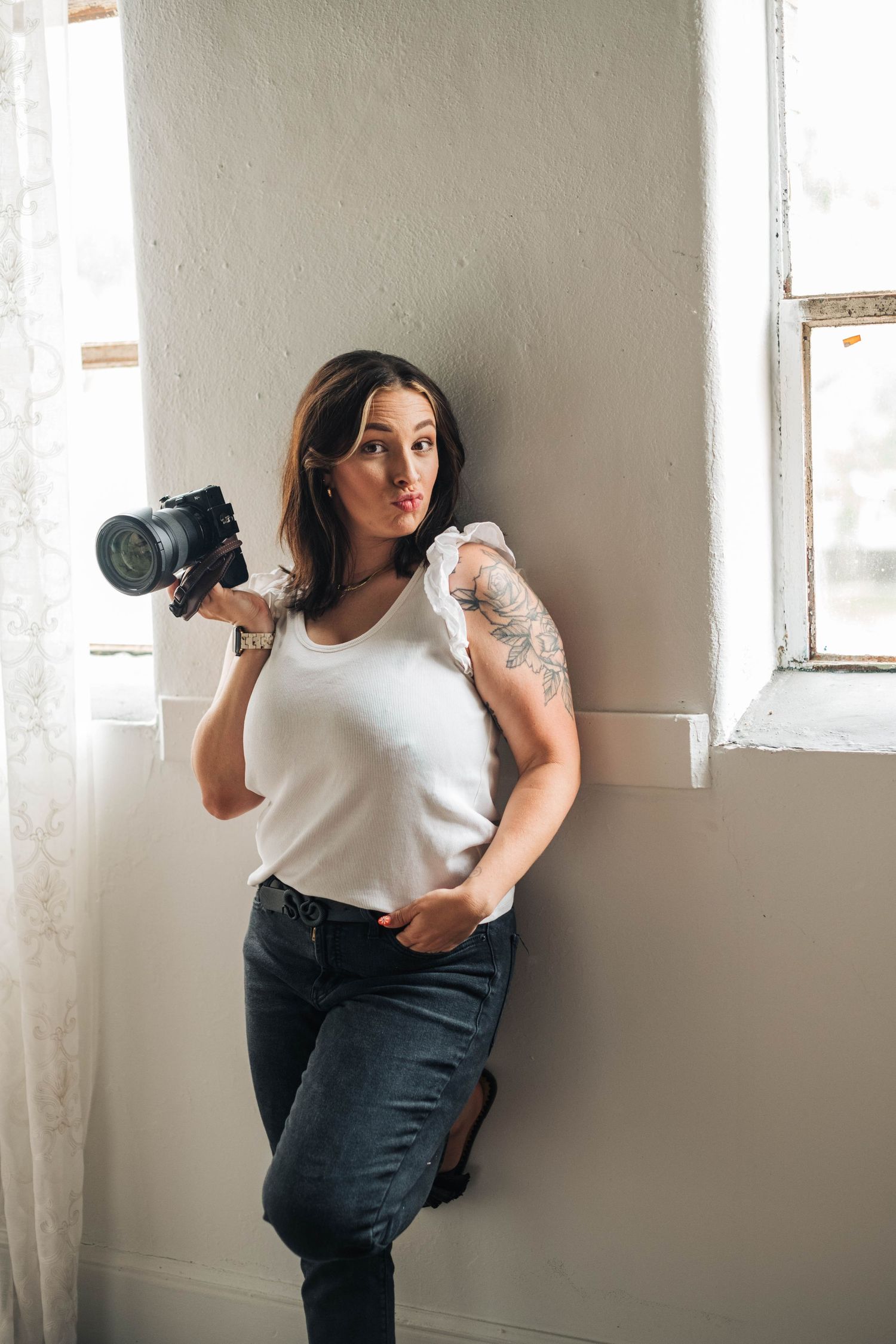 A photographer poses playfully while holding a camera in a white room during a creative self-portrait session.