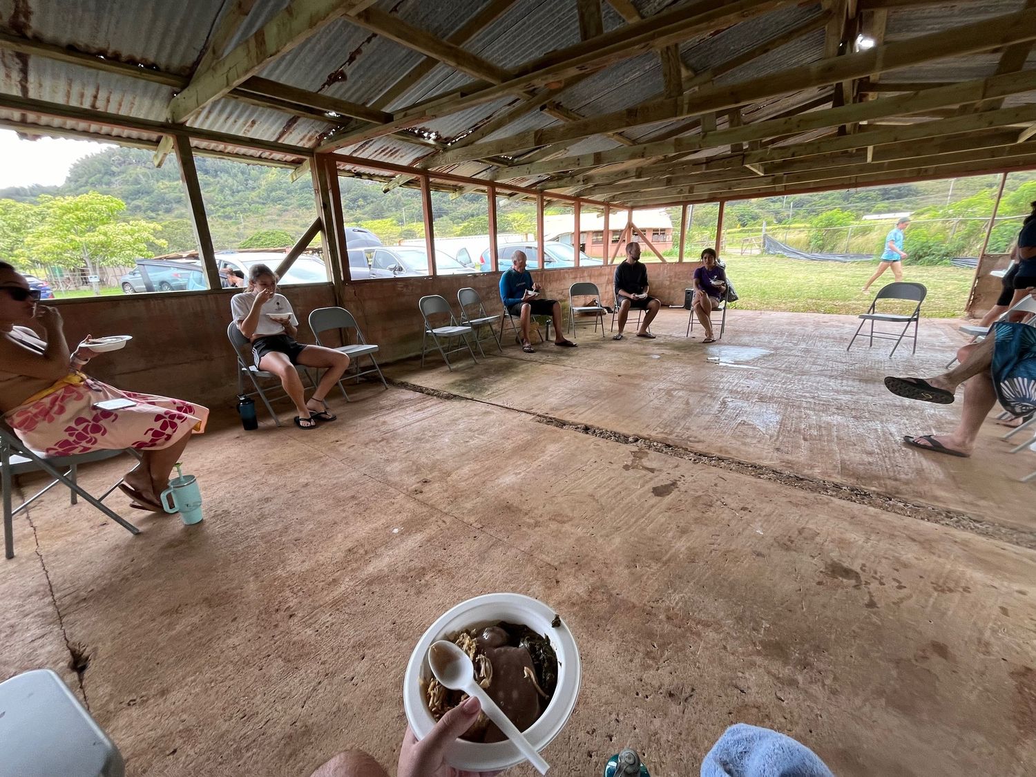 People gather in a rustic covered shelter with dirt floor and metal roof in a rural setting.