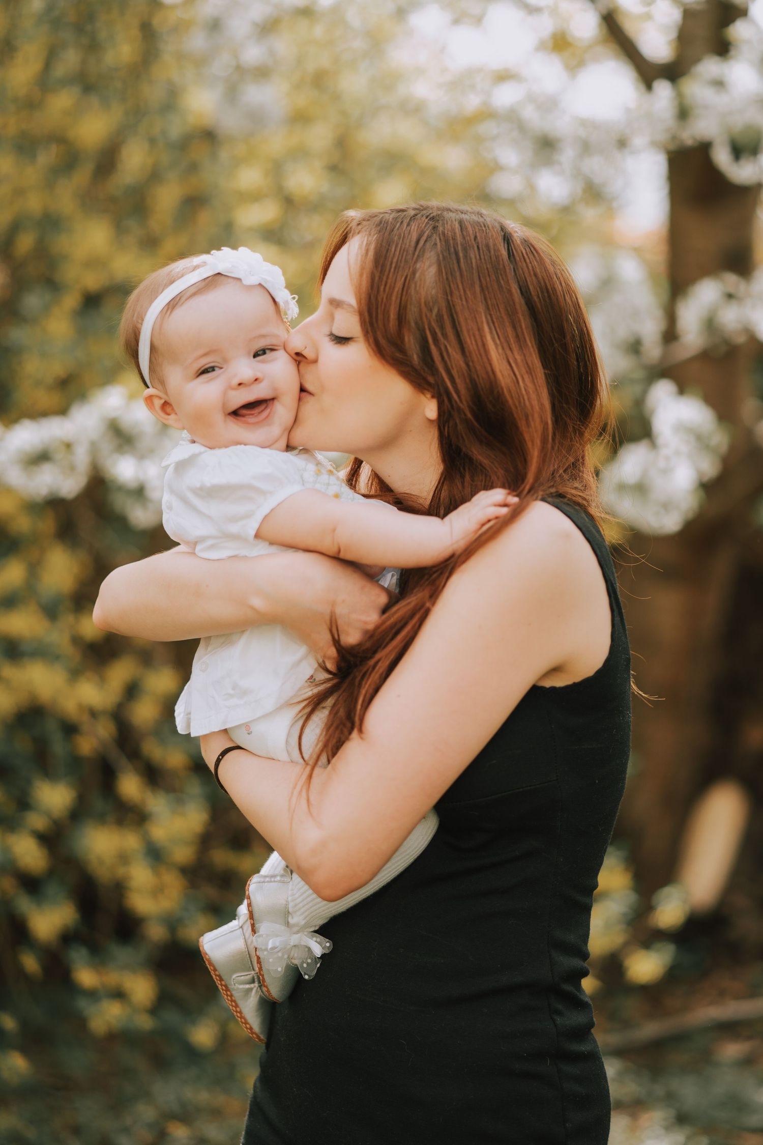 Mother in black tank top kisses baby wearing white outfit against natural backdrop.