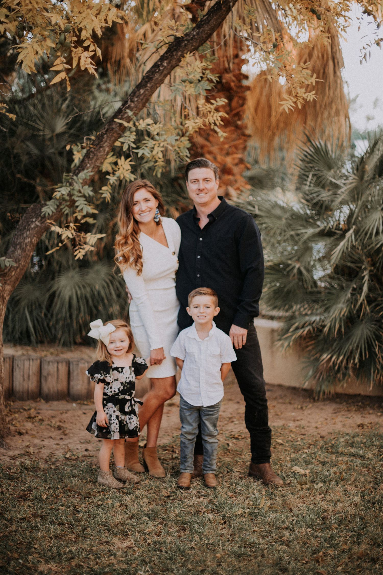A family poses together outdoors beneath autumn trees and palm fronds in warm golden lighting.