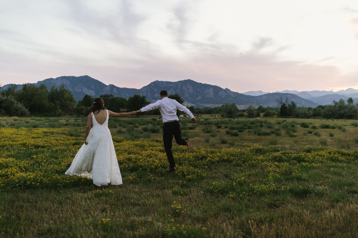Wedding couple runs through a field of yellow flowers with mountains in the background at sunset.